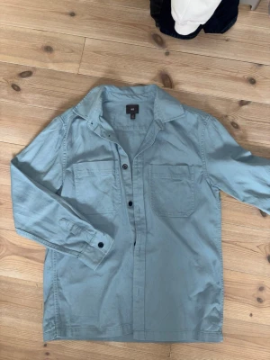 Ljusblå overshirt från H&M - Säljer en ljusblå skjorta från H&M i storlek S. Skjortan har klassisk krage, två bröstfickor och knappar framtill. Tillverkad i mjuk bomull och har långa ärmar med knapp vid manschetten. Perfekt för en avslappnad men snygg stil.
