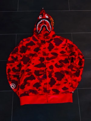 Röd camo hoodie från BAPE - Cool röd hoodie från BAPE med camouflagemönster och ikonisk hajdesign på huvan. Hoodien har dragkedja framtill och ribbade muddar. Perfekt för dig som vill sticka ut med streetwear-stil.