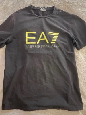 Svart EA7 Emporio Armani t-shirt - Cool svart t-shirt från EA7 Emporio Armani med stort gult tryck på bröstet. Klassisk rund hals och korta ärmar. Perfekt för dig som gillar stilrena och trendiga plagg med tydlig logga. Materialet är mjuk bomull som känns skönt mot huden.