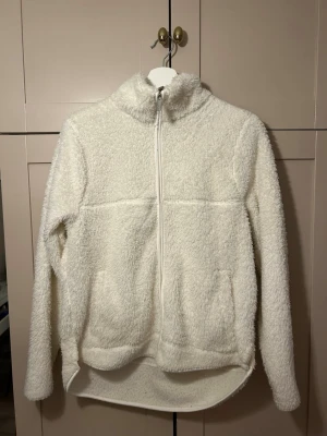 Vit teddyjacka från 157 - Mysig vit teddyjacka med dragkedja och hög krage från 157. Jackan har två stora fickor framtill och är tillverkad i fluffigt teddy-material. Perfekt för att hålla sig varm under kyliga dagar. Långärmad och något längre baktill för extra komfort.