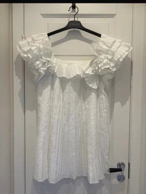 Vit plisserad klänning - Söt vit kortklänning med plisserad kjol och volangdetaljer vid axlar och halsringning. Klänningen är ärmlös och har en luftig passform, perfekt för studentfirande eller sommarens festligheter. Säljer denna då den är för liten! I super bra skick! Skicka gärna prisförslag!!!
