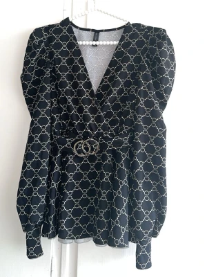 Svart tröja med bälte och mönster - Svart tröja med vitt cirkelmönster och markerad midja med snyggt bälte med dubbelring-detalj. Djup v-ringning och långa ärmar ger en trendig look. Perfekt för dig som gillar stilrena och moderna klänningar.