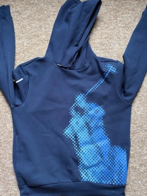 Mörkblå hoodie från Ralph Lauren - Snygg mörkblå hoodie från Ralph Lauren med stort blått pixelmönster av en polospelare på framsidan och vit signaturtext längs ena ärmen. Hoodien har huva och ribbade muddar. Perfekt för en avslappnad och cool stil.