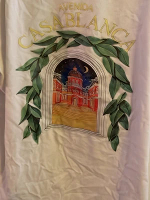 Vit t-shirt med Casablanca-tryck - Vit t-shirt från Casablanca med stort färgglatt tryck framtill. Trycket föreställer en byggnad med nattmotiv, omgiven av gröna blad och texten 'Avenida Casablanca' i gult. Klassisk passform och rund hals.