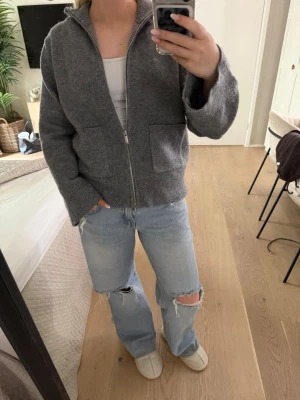 Grå ziptröja från Gina Tricot - Mysig grå ziptröja från Gina Tricot med två stora fickor framtill och hög krage. Tröjan är stickad i mjukt material och har långa ärmar. Perfekt för lager på lager och chill vardagsstil.