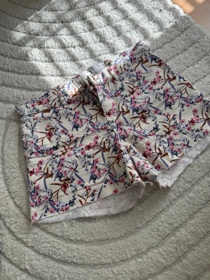 Blommiga shorts från H&M - Snygga blommiga shorts från H&M i vitt med rosa, lila och blått blommönster. Modellen är midwaist med råa kanter nertill och tillverkad i bomull. Perfekta för varma sommardagar och ger en fräsch och trendig look.