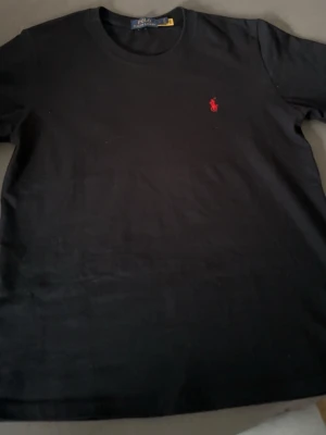 Polo Ralph Lauren - Klassisk svart t-shirt från Polo Ralph Lauren med rund halsringning och ikonisk röd broderad logga på bröstet. Tillverkad i mjuk bomull för en skön känsla och enkel stil. Perfekt basplagg för alla tillfällen.