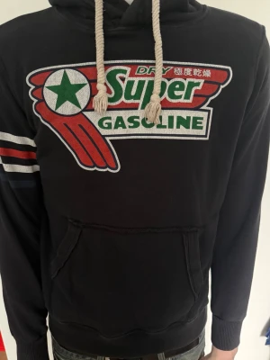 Svart hoodie Super Gasoline tryck - Svart hoodie med stort färgglatt Super Gasoline-tryck på bröstet och röda, vita och blå ränder på ena ärmen. Huva med grova vita snören och känguruficka framtill. Perfekt för dig som gillar retrostil och streetwear.