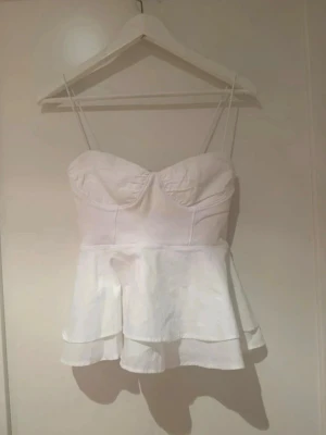 Vit topp - Supersöt vit topp med peplumvolang och tunna axelband. Toppen har markerade kupor och figurnära passform upptill. Helt ny med prislapp och aldrig använd.Liten i storlek!!💕