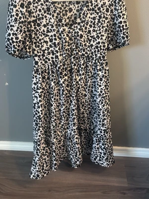 Leopardmönstrad klänning från Pimkie - Snygg leopardmönstrad klänning från Pimkie med kort ärm och knappar framtill. Klänningen har v-ringning och en luftig passform som ger en avslappnad vibe. Perfekt för dig som gillar att sticka ut med djurmönster.
