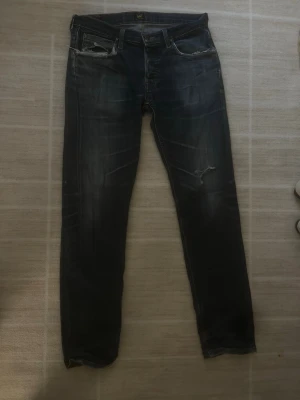 Vintage Mörkblå jeans från Lee - Säljer ett par mörkblå jeans från Lee med baggy fit och slitningar vid fickorna samt ett mindre hål på låret. Jeansen har klassisk femficksmodell och är tillverkade i denim med snygg tvätt och slitna detaljer. Perfekt för vintage och streetwear look