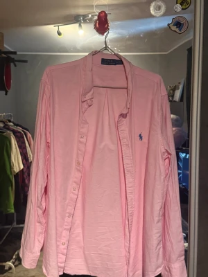 Rosa skjorta från Polo Ralph Lauren - Klassisk rosa skjorta från Polo Ralph Lauren med lång ärm och blå broderad logga på bröstet. Skjortan har knappar framtill och är tillverkad i mjuk bomull. Perfekt för en clean och stilren look.