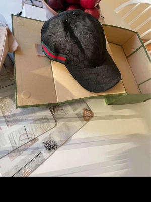 Svart Gucci keps med GG-mönster - Snygg svart keps från Gucci med det klassiska GG-monogrammet i tyget. Kepsen har gröna och röda ränder på sidorna som ger den en ikonisk look. Tillverkad i ett glansigt tyg som känns lyxigt och trendigt. Perfekt accessoar för att lyfta din outfit. Låda, påse och allt som ska följer med. 
