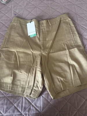 Beige shorts från The Glory Days - Snygga beige shorts från The Glory Days i rak modell med midja och knappstängning. Tillverkade i ett mjukt och svalt material som passar perfekt för varma dagar. Klassisk design med sidofickor och diskret look.