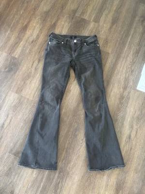 Grå bootcut jeans True Religion - Säljer ett par grå bootcut jeans från True Religion med klassiska fickor och silverfärgade knappar. Jeansen har låg midja och utsvängda ben, med snygga detaljer på bakfickorna. Stolek 28, modell joey, knappt använda🥰