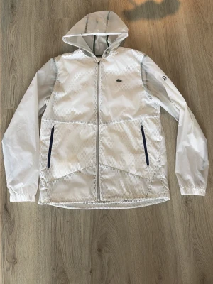 Vit vindjacka från Lacoste med huva - Snygg vit vindjacka från Lacoste med hel dragkedja, två svarta dragkedjefickor och justerbar huva. Jackan har diskret Lacoste-logga på bröstet och elastiska muddar vid ärmsluten. Materialet är lätt och vattenavvisande, perfekt för blåsiga dagar. Skick 10/10 bara köpt men aldrig använd.