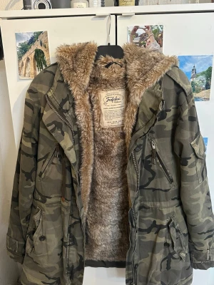 Kamo parkas med fuskpäls från Zara - Säljer en grön kamouflagemönstrad parkas från Zara med stor huva och mjukt fuskpälsfoder. Jackan har flera fickor med knappar och dragkedjor. Perfekt för kalla dagar och riktigt snygg streetstil. Är storlek XS men passar mig som ofta bär S.