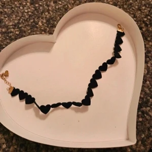 Svart sammetschoker med hjärtan - Chokerhalsband med svarta sammetsklädda hjärtan på rad och guldfärgade metalldetaljer. Perfekt accessoar för att ge din outfit en edgy och romantisk touch. Hjärtana är mjuka och halsbandet har ett klassiskt lås.
