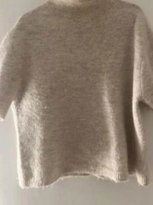 Beige stickad tröja med kort ärm - Mysig beige stickad tröja med korta ärmar och rund hals. Tröjan har en avslappnad passform och är gjord i ett fluffigt, mjukt material som känns skönt mot huden. Perfekt för lager-på-lager och enkel att matcha med olika outfits.