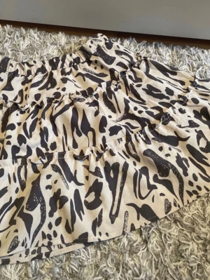 Kort kjol med leopardmönster - Snygg kort kjol med beige botten och svart leopardmönster. Kjolen har volangdetaljer och resår i midjan för en bekväm passform. Perfekt för dig som vill sticka ut med ett trendigt djurmönster.