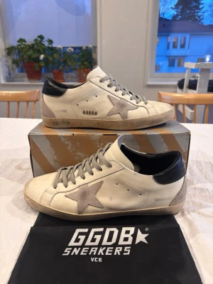 GGDB Superstar vita sneakers - Säljer ett par GGDB Superstar sneakers i vitt läder med grå mocka stjärndetalj på sidan, grå mocka vid hälen samt svart läderdetalj bak. Skorna har grå skosnören och klassisk låg silhuett. Perfekta för dig som gillar streetstyle och unika detaljer. Priset är ej hugget i sten! Endast använda i runt 3-4 veckor så endast lite smuts på de som man kan tvätta bort, har endast inte energin.