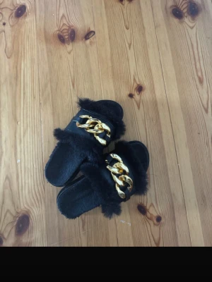 Svarta sandaler med päls och guldkedja - Coola svarta sandaler med mjuk fuskpäls och en stor guldfärgad kedjedetalj över foten. Sandalerna har en bred rem och räfflad sula för extra grepp. Perfekta för dig som vill sticka ut med din stil.
