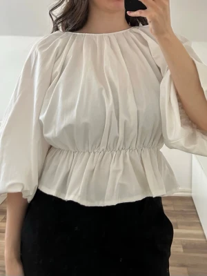 Vit puffärmad blus med resårmidja - Säljer en vit blus med puffiga ärmar och resår i midjan som ger en snygg siluett. Blusen har rund halsringning. Plagget var från början längre i modellen, men har sytts upp till en kortare och  smickrande modell. Storlek S. Aldrig använd. Material: 57% bomull och 43% polyester.