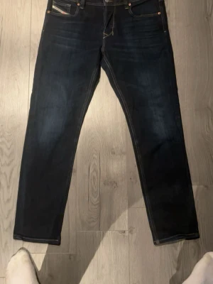 Mörkblå jeans från Diesel - Snygga mörkblå jeans från Diesel med klassisk femficksdesign och kontrastsömmar. Jeansen har en rak passform och detaljerad logga på myntfickan. Perfekta för dig som gillar stilrena och tidlösa jeans i slitstarkt denimtyg.