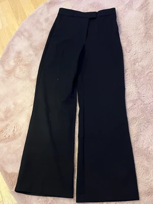 Svarta kostymbyxor från H&M - Snygga svarta kostymbyxor från H&M i klassisk rak modell med pressveck. Byxorna har hög midja och vida ben, perfekta för en stilren look. Materialet är mjukt och följsamt, vilket gör dem bekväma att bära hela dagen.