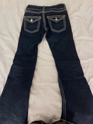 Mörkblå bootcut jeans med kontrastsömmar - Snygga mörkblå bootcut jeans med vita kontrastsömmar och dekorativa fickor med silverknappar. Jeansen har klassisk femficksdesign och orange detaljer vid sömmarna. Perfekt för dig som gillar en retrostil och vill sticka ut.