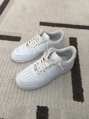 Vita Nike Air Force 1 sneakers - Klassiska helvita Nike Air Force 1 sneakers i lågt utförande. Skorna har ovandel i slätt läder, perforerade detaljer på tån och vit sula. Ikonisk Swoosh-logga på sidan och vita skosnören. Perfekta för en clean och stilren look.