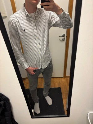 Vit, svart  slim fit skjorta från Ralph Lauren - Vit, svart randig skjorta med svarta vertikala ränder från Ralph Lauren. Slim fit-modell i 100% bomull med stretch, klassisk krage och knappstängning. Diskret logotyp broderad på bröstet. Perfekt för dig som gillar stilrena och snygga skjortor.