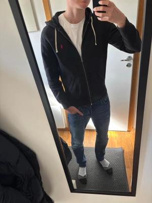 Svart hoodie från Polo Ralph Lauren - Säljer en svart hoodie från Polo Ralph Lauren med dragkedja, vit dragsko och klassisk röd logga på bröstet. Hoodien har fickor framtill och en skön huva. Perfekt för en avslappnad och stilren look.