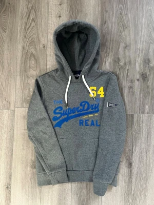 Grå Superdry hoodie med tryck - Snygg grå hoodie från Superdry med blått och gult tryck framtill och vita dragsnören i huvan🤩 passar nån i storlek Xs och inga fel😇 hör av er vid snabbaffär samt funderingar
