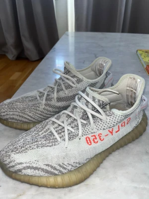 Yeezy Boost 350 V2 Zebra sneakers - Säljer ett par Adidas Yeezy Boost 350 V2 Zebra med stickad ovandel i vitt och grått mönster, röda SPLY-350-detaljer på sidan och en genomskinlig, beige sula. Skorna har en låg silhuett och klassiska vita snören.