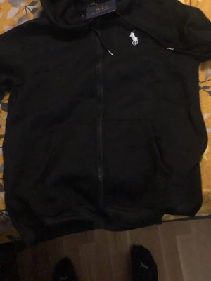 Svart hoodie från Polo Ralph Lauren - Svart hoodie med dragkedja från Polo Ralph Lauren. Klassisk design med vit broderad logga på bröstet, huva med dragsko och stora fickor framtill. Perfekt för en chill och stilren look.