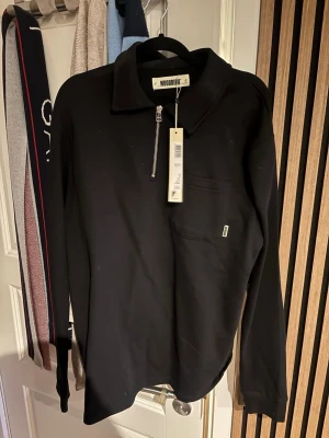 Svart långärmad tröja Woodbird med prislapp! - Svart långärmad tröja från Woodbird med halv dragkedja och krage. Tröjan har en bröstficka med liten logotypdetalj och är tillverkad i ett mjukt material. Ny med prislapp!