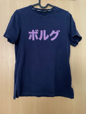 Mörkblå t-shirt från Björn Borg - Mörkblå t-shirt från Björn Borg med lila japansk text framtill och 'BORG' tryckt i nacken. Klassisk rund hals och korta ärmar. Tillverkad i mjuk bomull för en skön känsla och enkel stil.