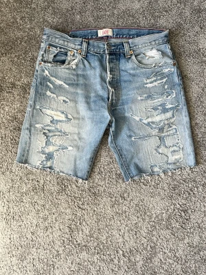Levi's 501  jeansshorts - Ljusblå jeansshorts från Levi's 501 med rejält slitna detaljer och råa kanter. Klassisk femficksmodell med knappgylf och avslappnad passform. Perfekta för en avslappnad streetstil under varma dagar. Bra skick. Storlek är 34 men sitter som 32 i midjan. Säljes då jag växt ut dem. Pris 299