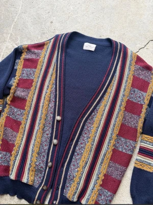 Cardigan 80tal  - Retro kofta i mörkblått med färgglada ränder och mönster i vinrött, gult, blått och grått. Djup V-ringning och knäppning med fyra metallknappar. Stickad i mjukt material, perfekt för lager-på-lager och unik stil.