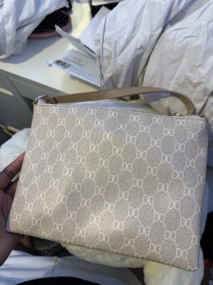 Beige kuvertväska från Don Donna - Stilren beige kuvertväska från Don Donna med monogrammönster i vitt.