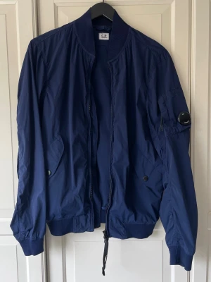 C.P. Company bomberjacka sommarjacka - En lätt jacka i klassisk bomberjacka-stil från legendariska CP Company. Perfekt för vår, sommar och till höst med en hoodie under.  Hela jackan är i fint skick förutom en 1 cm repa på vänster ärm (se bild). Knappt märkbar.  Passar mig bra; jag är 176 cm och väger 70 kg.  Axel: 46 cm Arm: 66 cm Rygg: 70 cm