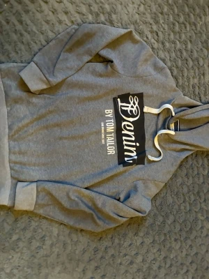 Grå hoodie Tom Tailor Denim - Säljer en grå hoodie från Tom Tailor Denim med stor vit och svart logga på bröstet. Hoodien har huva med vita dragsnören och ribbade muddar vid ärmar och nederkant. Perfekt för en avslappnad och stilren look.