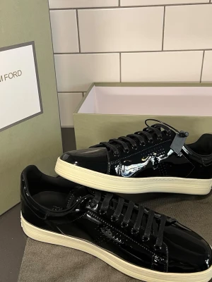  Tom Ford skor - Snygga svarta sneakers från Tom Ford i glansigt lackmaterial med vita sulor. Klassisk låg modell med svarta skosnören och diskret perforerat mönster på sidan. Perfekta för dig som vill sticka ut med stilrena och exklusiva skor.