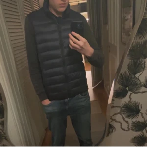 Svart dunväst med dragkedja - Snygg svart dunväst med horisontella sömmar och hög krage. Västen har dragkedja framtill och två sidofickor. Perfekt att slänga över en hoodie eller tröja för extra värme. Enkel och stilren design som passar till det mesta.