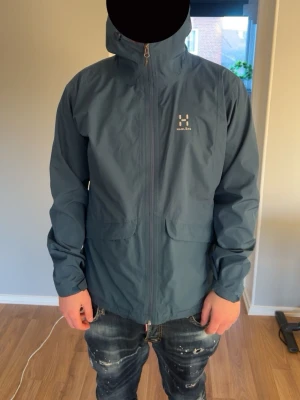 Blå vindjacka från Haglöfs med huva - Snygg blå vindjacka från Haglöfs med justerbar huva och dragkedja framtill. Jackan har två stora fickor med lock och diskret Haglöfs-logga på bröstet. Tillverkad i lätt och slitstarkt material, perfekt för blåsiga dagar. Xl för tjejer o storlek l för killar 