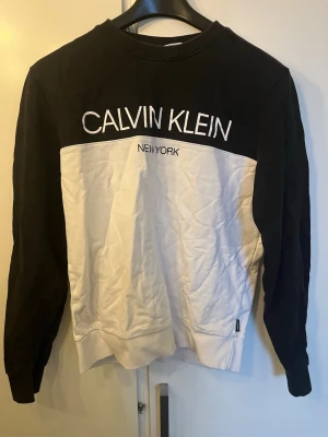 Svartvit sweatshirt från Calvin Klein - Snygg svartvit sweatshirt från Calvin Klein med stort tryck på bröstet där det står 'CALVIN KLEIN NEW YORK'. Tröjan har rund halsringning, långa ärmar och är tillverkad i mjukt bomullsmaterial. Perfekt för en avslappnad och trendig look.