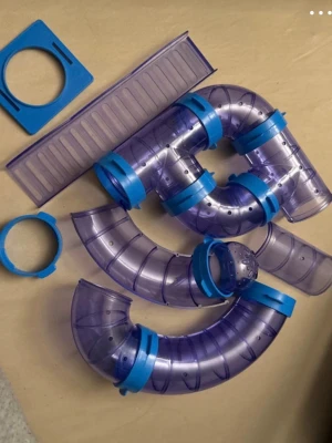 Hamsterhjul och tunnelsystem plast - Säljer ett set med ett orange hamsterhjul och flera lila-blåa plasttunnlar med kopplingar, perfekt för smådjur som hamster eller gerbil. Tunnlarna är böjda och kan byggas ihop på olika sätt. Allt är gjort i tålig plast.