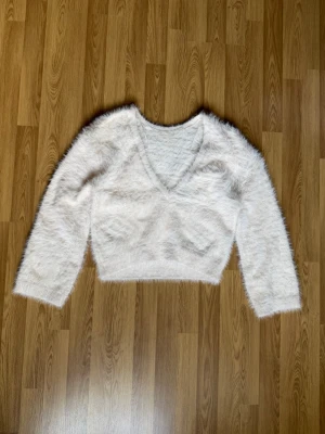 Vit fluffig stickad tröja - Supermjuk och fluffig vit stickad tröja med djup v-ringning och långa ärmar. Tröjan har en croppad passform och är perfekt för att skapa en mysig och trendig look. Materialet är lurvigt och ger en cool textur.