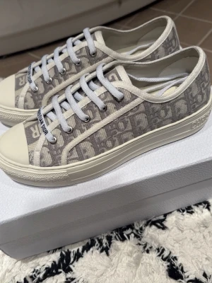 Grå Dior sneakers med monogram - Snygga sneakers från Dior i grått och vitt med klassiskt Dior-monogram över hela skon. Skorna har vita snören, gummisula och detaljer med 'CHRISTIAN DIOR' på sidan och tungan. Tillverkade i textil och gummi för en trendig och exklusiv look.